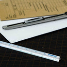 Een Penco Lat Aluminium Zilver, een hobbymes en vellen papier, waaronder een bruin formulier, liggen netjes op een donkere snijmat met maatlijnen - ideaal voor technische tekenklussen.