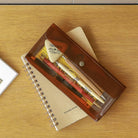 De Nähe Multifunctionele (Pennen)Zak Bruin van Nähe, gevuld met pennen, potloden en een gum, ligt op een spiraalgebonden notitieblok met college-blad op een houten bureau.