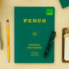 Een Penco Notitieboek Groen A5 met een pen, een geel potlood met rode gum en een klein klembord met papier liggen netjes gerangschikt op een licht houten oppervlak.