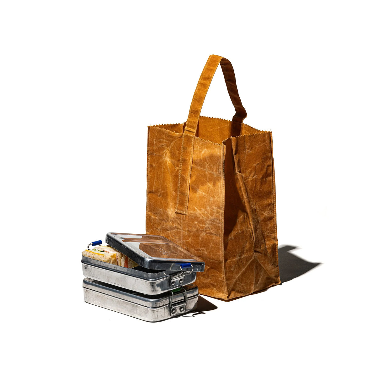 Een bruine herbruikbare papieren tas met hengsels staat rechtop naast twee gestapelde aluminium lunchboxen van Puebco, gevuld met eten, tegen een effen witte achtergrond.