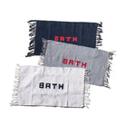 Drie Puebco Badmat Gerecycleerd Garen Blauw 75/45 matten, handgeweven in marineblauw, grijs en wit met franjeranden en "BATH" erop geweven, liggen verspringend op een witte achtergrond.