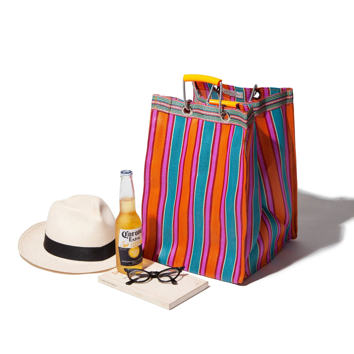 Een Puebco Gerecycleerde Tas Strepen Oranje Roze D26 zit naast een witte fedora hoed, een fles Corona bier, een zwarte bril en een paperback boek tegen een witte achtergrond.