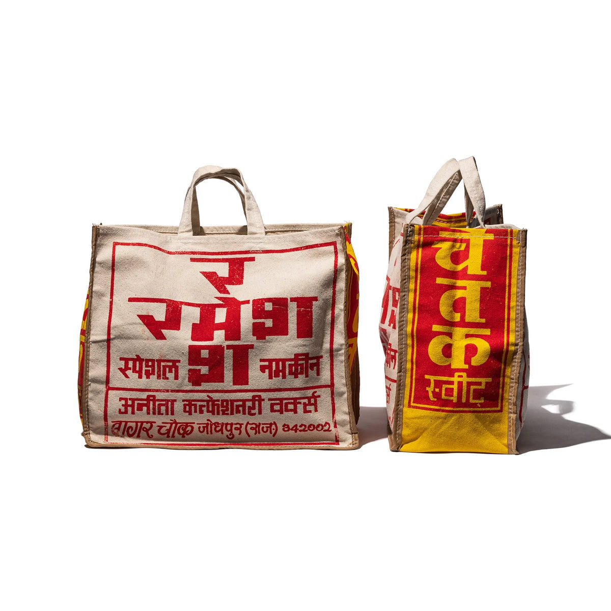Twee beige en rode Puebco India Souvenir Tasjes met vetgedrukte Hindi tekst in rood en geel staan rechtop - de ene naar voren gericht, de andere opzij gedraaid. Deze Puebco boodschappentassen zijn perfect als uniek souvenir of stijlvolle dagelijkse tas.