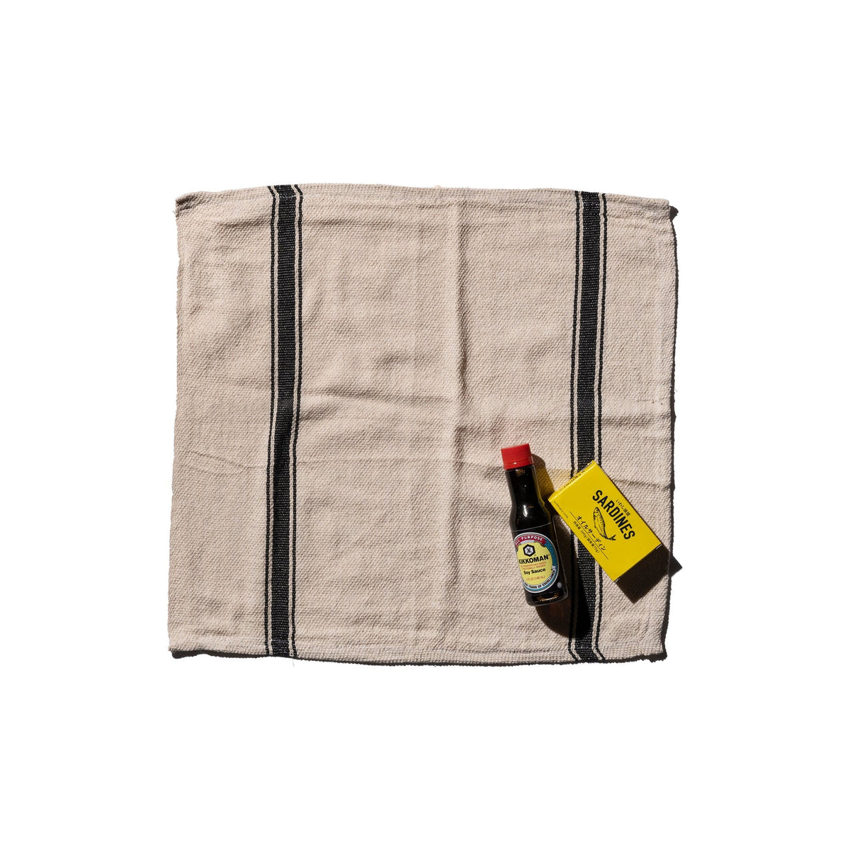 Een gevouwen Puebco India Theedoek Zwart 60/60 van Puebco, gemaakt van gerecycleerde stoffen met zwarte strepen, staat naast een klein flesje sojasaus en een geel Sabadiso doosje op een witte achtergrond.