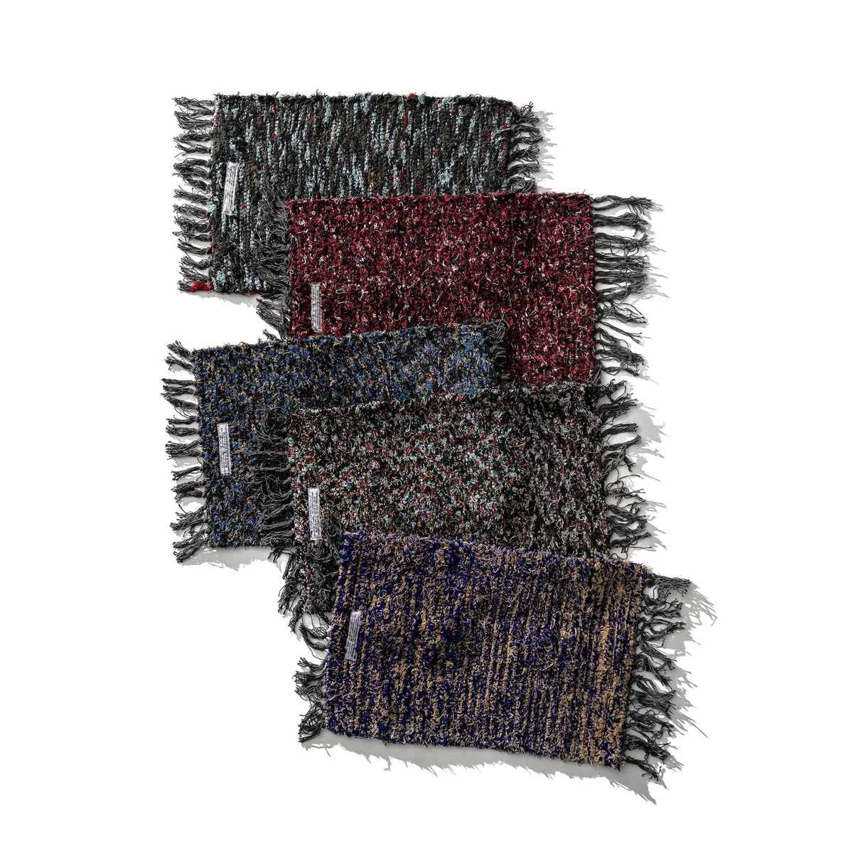 Vier rechthoekige Puebco Mat Gerecycleerd Textiel 63/41 geweven matten van Puebco, elk in zwart, rood, blauw en bruin met franjeranden en labels linksonder, liggen diagonaal over elkaar.