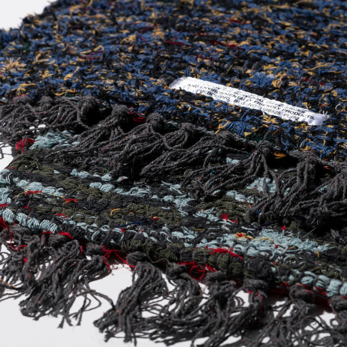 Een close-up van de Puebco Mat Gerecycleerd Textiel 63/41 van Puebco, met de veelkleurige geweven draden-blauw, zwart, rood, geel-omzoomde randen en een klein wit label met gedeeltelijk zichtbare tekst.