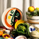 Op een tafel staan kleurrijke gerechten uitgestald, met in de hoofdrol het Serax Feast Ottolenghi Serveerbord Face N°1 D35 – een groot serveerbord van Serax, versierd met een abstract gezichtsontwerp geïnspireerd door Yotam Ottolenghi – omringd door kommen, mokken, groene kool, meloen, druiven, bessen en een fruitschaal, allemaal tegen een witte gordijnachtergrond.