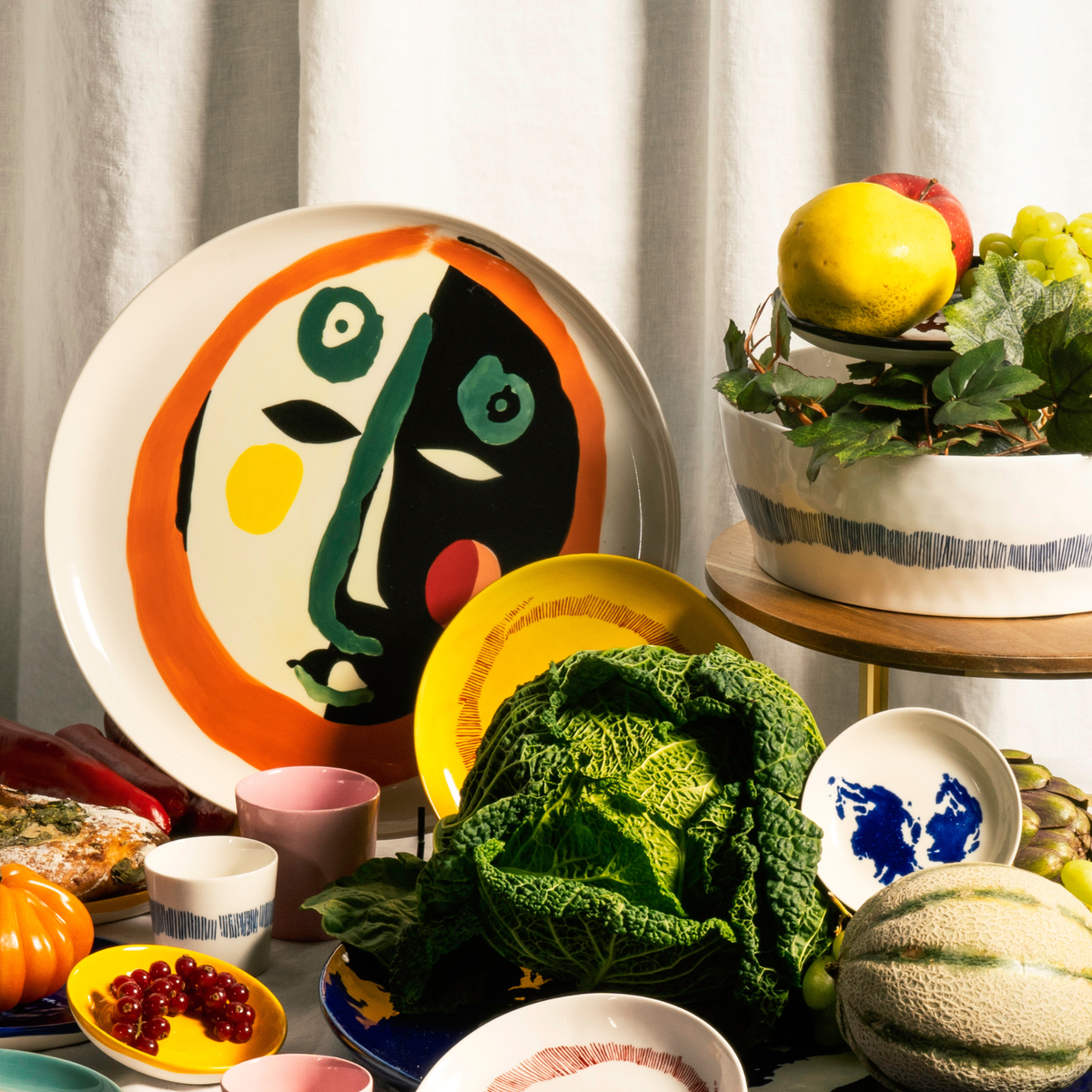 Op een tafel staan kleurrijke gerechten uitgestald, met in de hoofdrol het Serax Feast Ottolenghi Serveerbord Face N°1 D35 – een groot serveerbord van Serax, versierd met een abstract gezichtsontwerp geïnspireerd door Yotam Ottolenghi – omringd door kommen, mokken, groene kool, meloen, druiven, bessen en een fruitschaal, allemaal tegen een witte gordijnachtergrond.