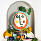 Een levendig assortiment van kleurrijke borden, kommen en kopjes is kunstig gerangschikt rond het Serax Feast Ottolenghi Serveerbord Face N°2 D35 van Serax – met zijn opvallende abstracte gezichtsontwerp geïnspireerd door Yotam Ottolenghi – omringd door vers fruit, bladgroenten en kruiden, allemaal geplaatst binnen een decoratieve witte boog.