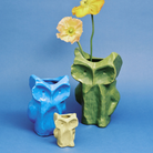 Drie Serax Marni After Midnight Uil Vaas Blauw S keramische uilenvazen in blauw, groen en lichtgeel staan op een blauw oppervlak; in de groene vaas zitten twee gele klaproosbloemen.