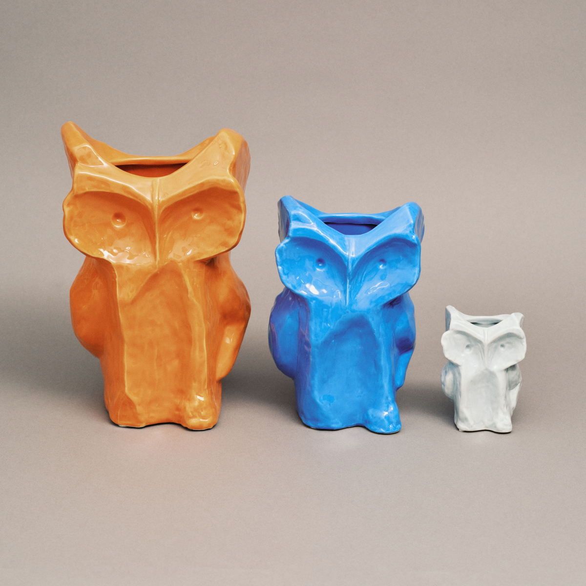 Drie keramische vazen in de vorm van een uil - waaronder een grote oranje vaas, een blauwe Serax Marni After Midnight Uil Vaas Blauw S en een kleine witte vaas - staan op een rij op een neutrale achtergrond.