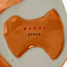 Close-up van de Serax Marni After Midnight Uil Vaas Oranje S, een vaas van keramiek met een glanzend oranje basis en "MARNI" in rood en "SERAX" in zwart op het gladde gebroken witte oppervlak.
