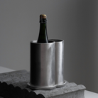 In de strakke, zilverkleurige Serax Surface Wijnkoeler van Serax, elegant vervaardigd uit verfijnd aluminium, staat een groene wijnfles met een kurk. De wijnkoeler staat op een grijs oppervlak met structuur, tegen een effen, zacht verlichte achtergrond.