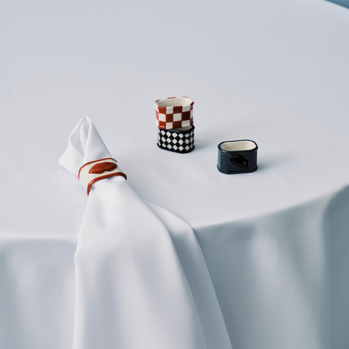 Een wit tafelkleed bedekt een ronde tafel. Bovenop ligt een gevouwen wit servet met een Serax Wouters & Hendrix servetring, een klein kopje met rode en zwarte motieven en een blauw vierkant bakje met een zwart deksel.