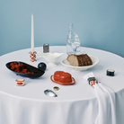 Een ronde witte tafel met brood, frambozen, een rood botervlootje, een kaars, een waterfles en een elegant decor - benadrukt door de Serax Wouters & Hendrix Servetring 4 st. tegen een lichtblauwe achtergrond.
