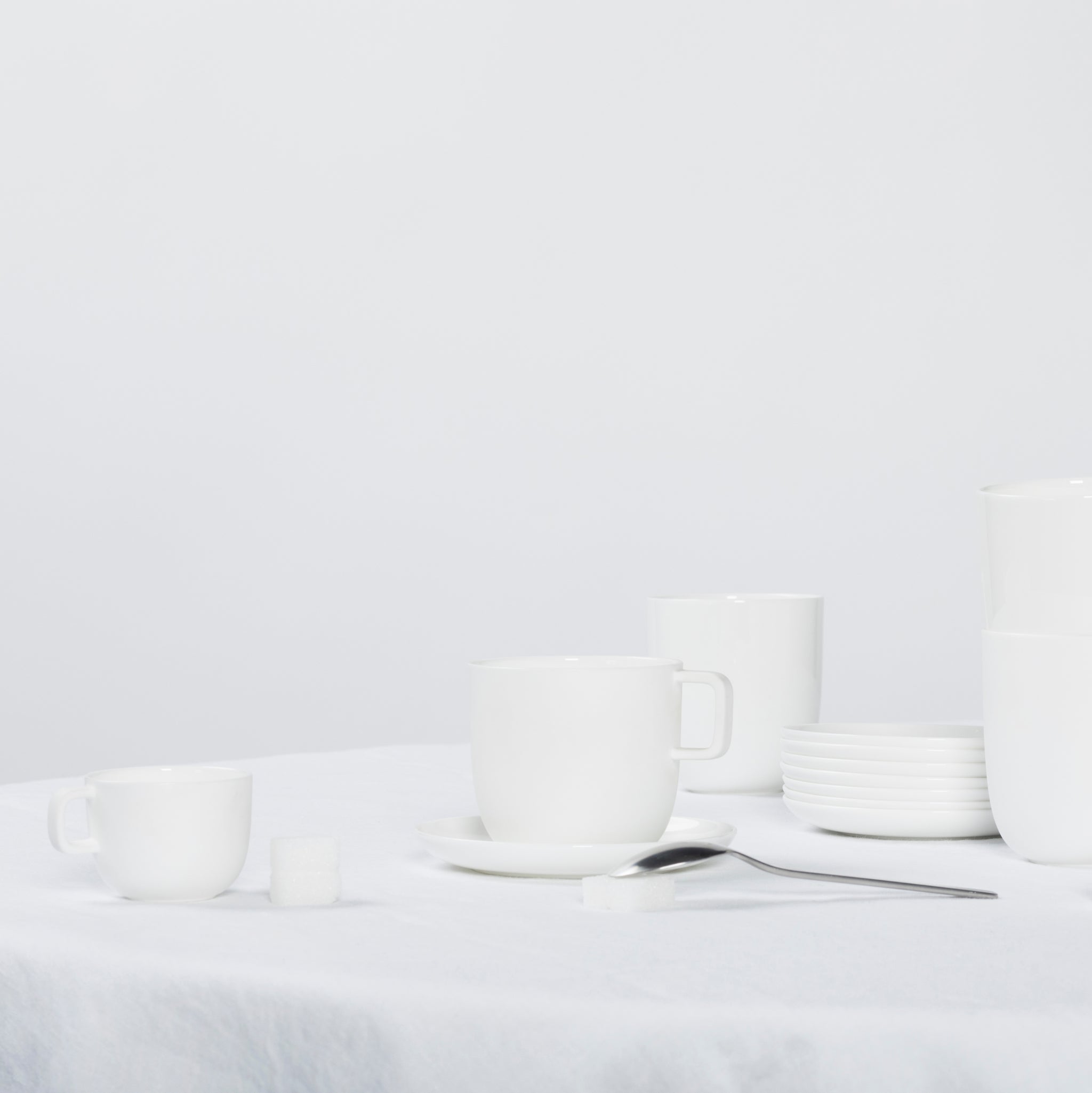 De Serax Base Koffiekop met Oor 30 cl van Serax is gerangschikt met bijpassende schotels en borden op een wit tafelkleed, naast een lepel en suikerklontjes; de volledig witte achtergrond creëert een strakke, minimalistische scène.