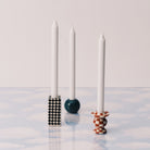 Drie witte kaarsen in unieke houders: de Serax Wouters & Hendrix Chessboard Kandelaars (set van 3) in zwart en wit, een blauwe bolhouder en een rood geruite voet - allemaal op een blauw en wit oppervlak met wolkenpatroon.