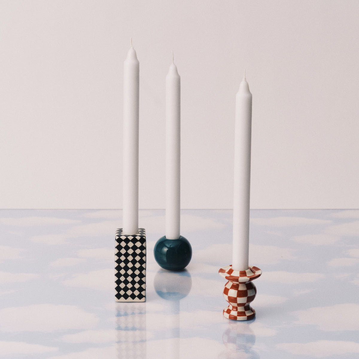 Drie witte kaarsen in unieke houders: de Serax Wouters & Hendrix Chessboard Kandelaars (set van 3) in zwart en wit, een blauwe bolhouder en een rood geruite voet - allemaal op een blauw en wit oppervlak met wolkenpatroon.