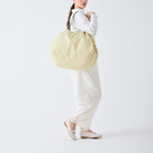 Een vrouw in witte kleding en loafers draagt een grote, beige Shupatto Opvouwbare Boodschappentas Cashew Nuts L over haar schouder, terwijl ze tegen een effen witte achtergrond staat.