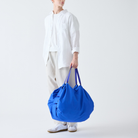 Een persoon in een los wit overhemd, een lichte broek en witte sneakers houdt een Shupatto Opvouwbare Boodschappentas Clear Sky Blauw L-draagtas met twee hengsels vast tegen een effen witte achtergrond.