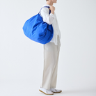 Een persoon in een wit overhemd en een lichte broek staat in profiel afgebeeld, met de Shupatto Opvouwbare Boodschappentas Clear Sky Blauw L over de schouder. Tegen een effen witte achtergrond draagt hij witte sneakers.
