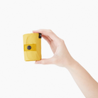 Op een effen witte achtergrond is een hand te zien die de Shupatto Opvouwbare Boodschappentas Mosterd M vasthoudt, een compacte gele opvouwbare boodschappentas van Shupatto met een drukknoopsluiting.