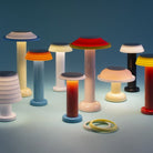Een groep kleurrijke, moderne Sowden PL1 Lamp LED Oplaadbaar Zwart lampen van Sowden, met cilindrische lampvoeten en gelaagde schijfvormige kappen geïnspireerd door de Memphis Design Movement, zijn gerangschikt op een blauw oppervlak met twee ronde ringen op de voorgrond.