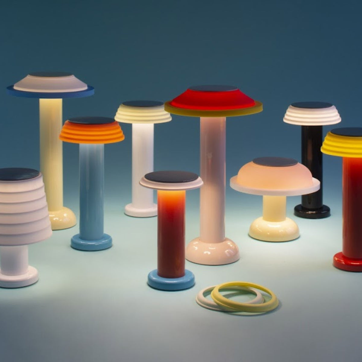 Een groep kleurrijke, moderne Sowden tafellampen, waaronder de Sowden PL2 Lamp LED Oplaadbaar Geel, zijn gerangschikt op een blauw oppervlak met zachte verlichting. Twee cirkelvormige banden rusten in de rechter voorhoek tegen een blauwe achtergrond met kleurverloop.