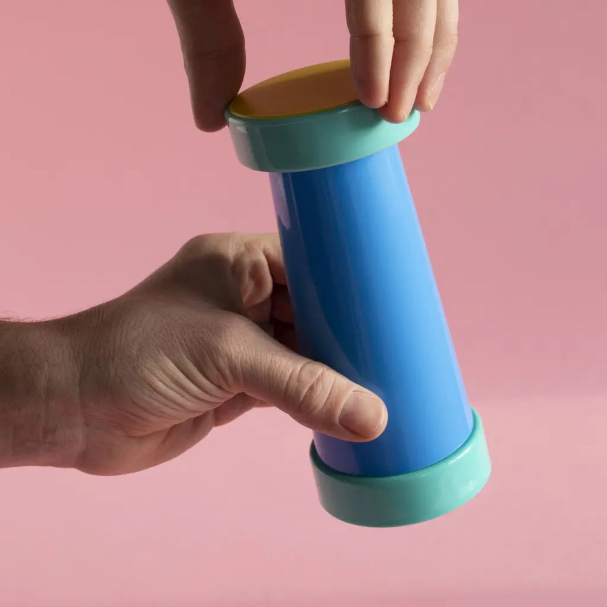 Twee handen houden de Sowden Zout-en Pepermolen Blauw van Sowden vast, een kleurrijke plastic zout- en pepermolen met een blauwe cilinder, groene uiteinden en een oranje bovenkant, geïnspireerd door de Memphis Design Movement, tegen een roze achtergrond.