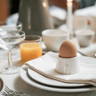 Een bruin ei rust in een Stock Antwerpen Eierdopje Wit Spikkel op een met servet bekleed bord, omringd door uniek servies, jus d'orange, een champagnecoupe, kopjes en een brandende kaars - ideaal voor het ontbijt van Kringwinkel Antwerpen.