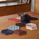 Een zwarte pan met deksel staat op een houten tafel, rustend op Zone Denmark Triangles Trivet Blauw siliconen onderzetters in verschillende kleuren en geometrische patronen. Op de achtergrond staan een houten kast en een raam.