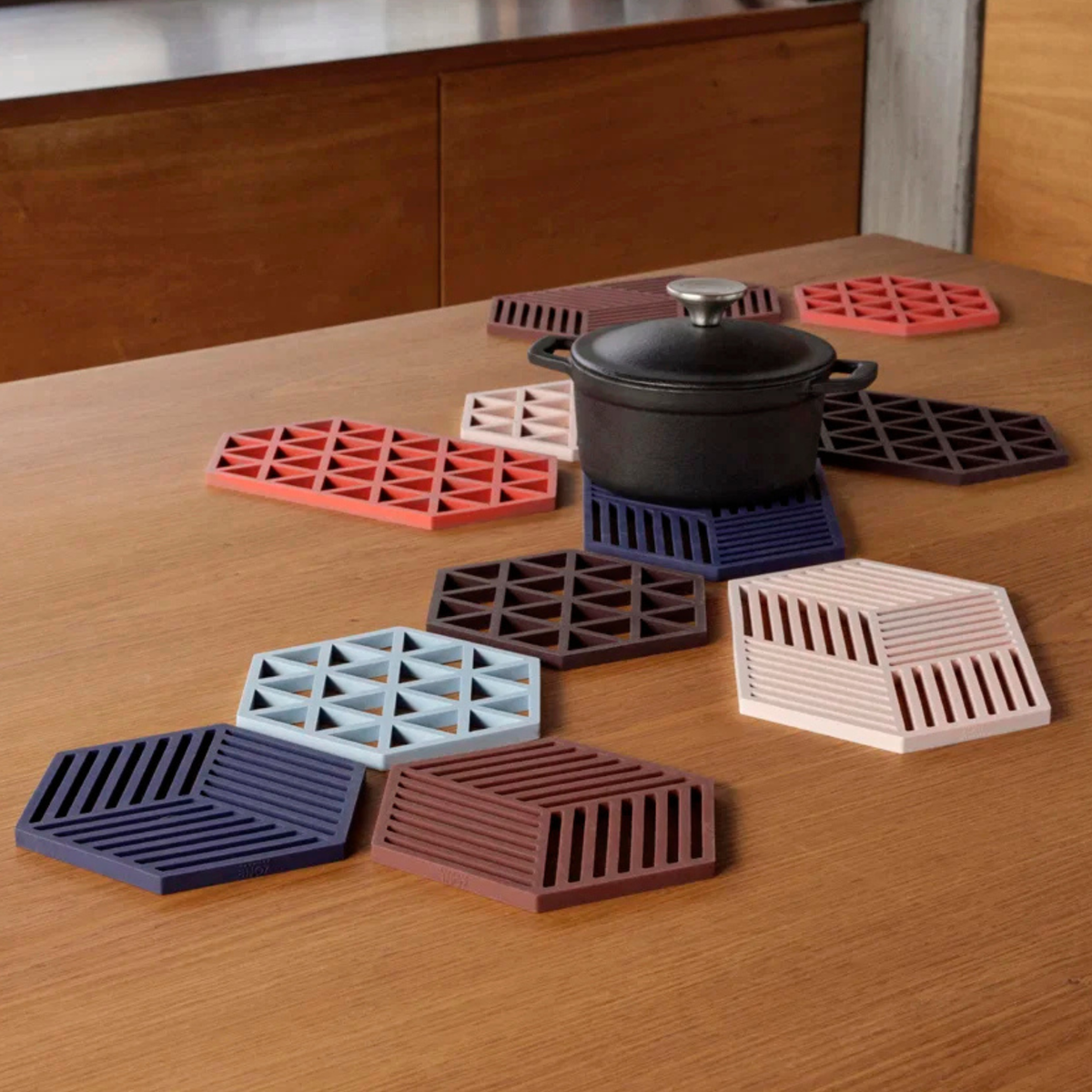 Op een houten tafel staat een zwarte pot, omringd door de Hexagon Trivet XL Roze van Zone Denmark en kleurrijke siliconen onderzetters, allemaal gerangschikt in een cirkelvormig geometrisch patroon.