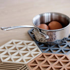 Een kleine roestvrijstalen steelpan met twee bruine eieren rust op een Zone Denmark Triangles Trivet Zwart van Zone Denmark op een licht houten oppervlak.