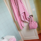 Een Bongusta Naram Badhanddoek Baby Pink & Ski Patrol 70/140 hangt aan een witte deur naast een bijpassende roze gestreepte tas. Licht blauwgroene tegels langs de muur, met een deel van een bad zichtbaar in de linker benedenhoek.