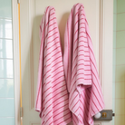 Twee Bongusta Naram badhanddoeken in Baby Pink & Ski Patrol hangen zij aan zij op haken aan een witte deur in de badkamer. Op de achtergrond zie je lichtgroene en witte tegels.