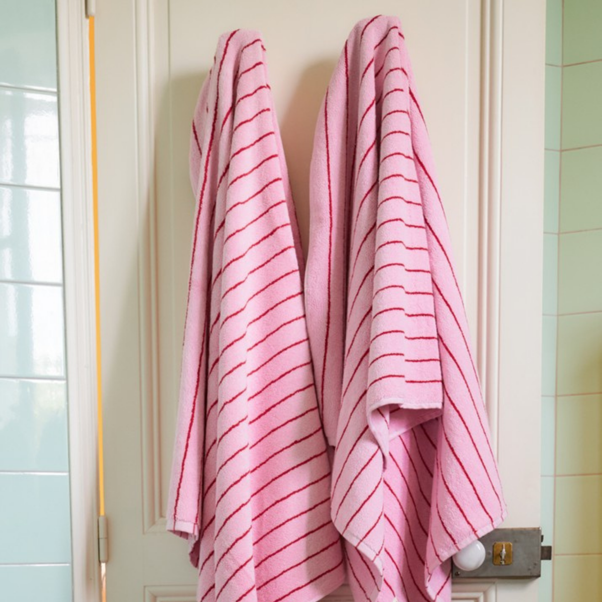 Twee Bongusta Naram badhanddoeken in Baby Pink & Ski Patrol hangen zij aan zij op haken aan een witte deur in de badkamer. Op de achtergrond zie je lichtgroene en witte tegels.
