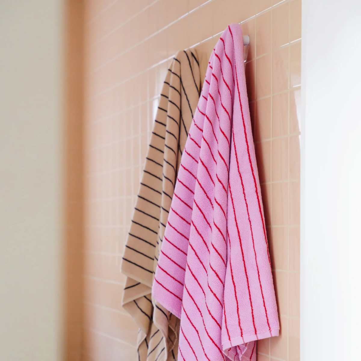 Twee Bongusta Naram Badhanddoeken - de ene in beige met zwarte strepen, de andere in babyroze met rode strepen - hangen naast elkaar aan een haakje tegen een licht perzikkleurig betegelde badkamermuur. Beide zijn gemaakt van biologisch katoen.