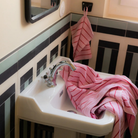 Een kleine witte wastafel in een betegelde badkamer heeft de Bongusta Naram Badhanddoek Baby Pink & Ski Patrol 70/140, gemaakt van biologisch katoen, over de wastafel gedrapeerd. Een andere soortgelijke Bongusta handdoek hangt erboven. Een spiegel en een raam zijn zichtbaar.