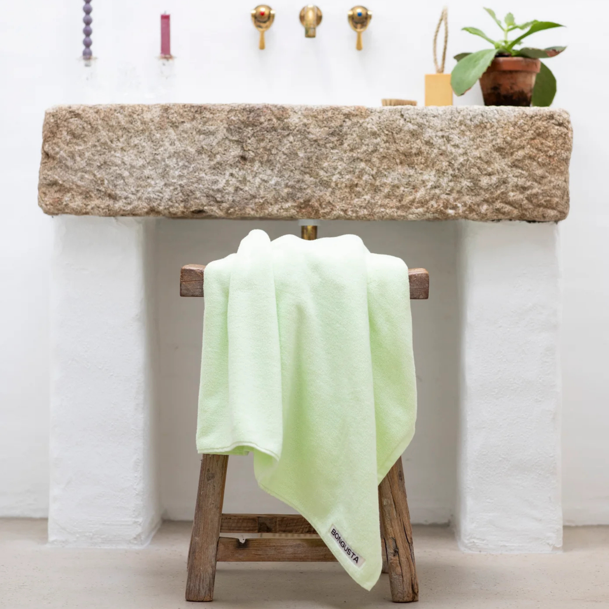 De Bongusta Naram Handdoek Lemon Sorbet 50/80 van Bongusta ligt over een rustiek houten krukje voor een stenen wastafel met drie messing kranen, kaarsen en een plantje op het witte aanrecht.