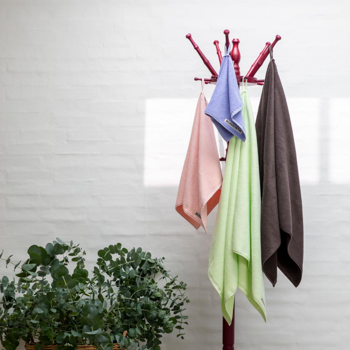 Een rode kapstok staat tegen een witte bakstenen muur met vier handdoeken in pastelroze, lavendel, lichtgroen en een Bongusta Naram Badhanddoek Lemon Sorbet 70/140. Linksonder staat een groene plant met bladeren.
