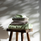 Een Bongusta Naram Badhanddoek Lemon Sorbet 70/140 ligt netjes opgevouwen in zachte groene en beige tinten op een rustiek houten krukje, zonlicht en schaduwen spelen over de witte muur erachter.