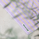 De Bongusta Naram Badmat Baby Lilac & Neon Yellow 50/80, gemaakt van biologisch katoen met dunne neongele strepen en een BONJOUR palmboomlabel, rust op een licht gespikkeld oppervlak met vage groene bladeren op de voorgrond.