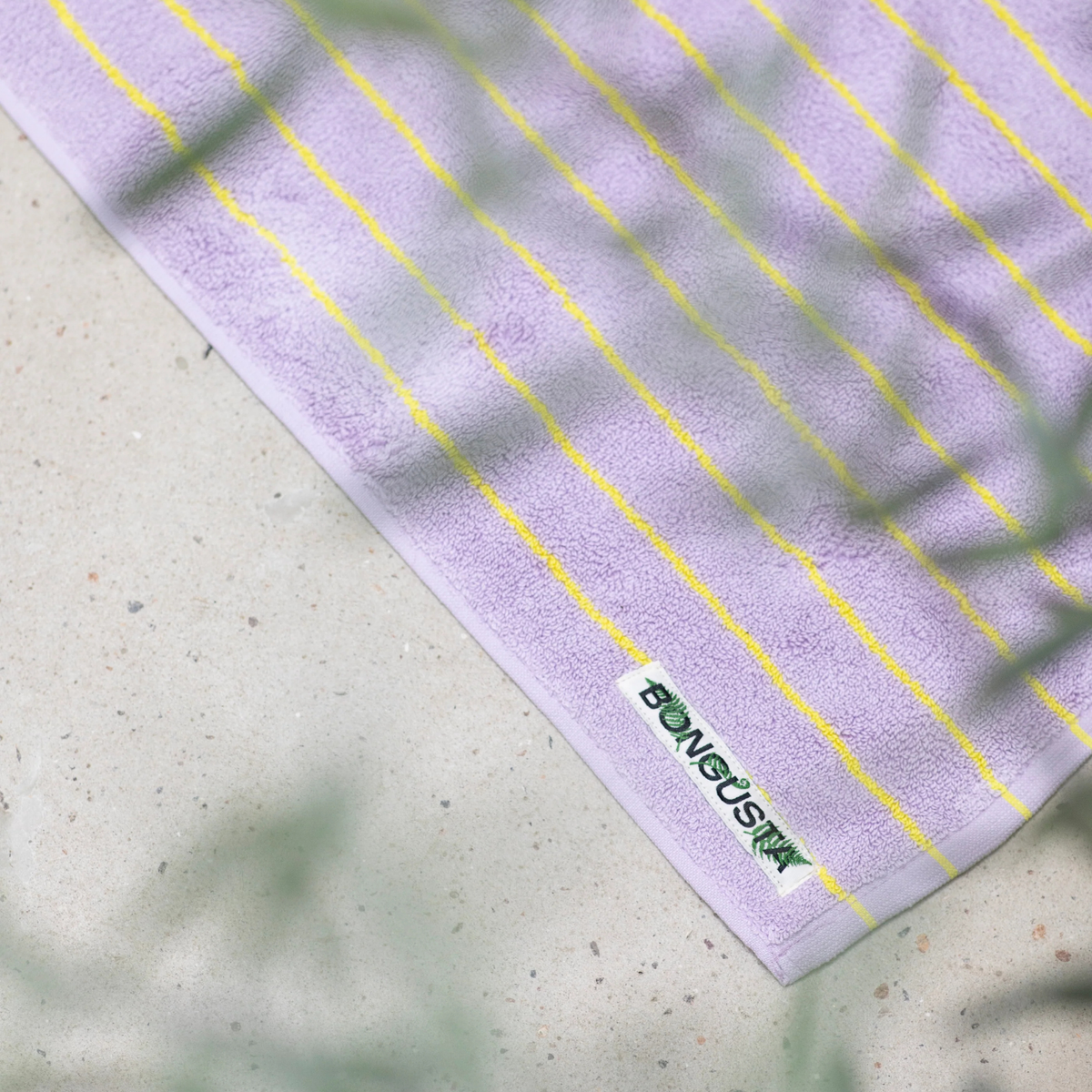 De Bongusta Naram Badmat Baby Lilac & Neon Yellow 50/80, gemaakt van biologisch katoen met dunne neongele strepen en een BONJOUR palmboomlabel, rust op een licht gespikkeld oppervlak met vage groene bladeren op de voorgrond.