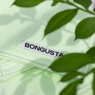 Een gevouwen lichtgroene Bongusta Naram Handdoek Lemon Sorbet 50/80, gemaakt van zacht biologisch katoen met een wit "BONGUSTA" label, gedeeltelijk bedekt door groene bladeren op de voorgrond.