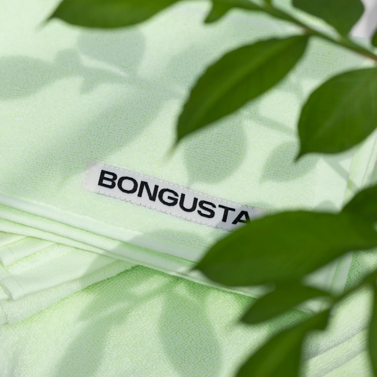 Een gevouwen lichtgroene Bongusta Naram Handdoek Lemon Sorbet 50/80, gemaakt van zacht biologisch katoen met een wit "BONGUSTA" label, gedeeltelijk bedekt door groene bladeren op de voorgrond.