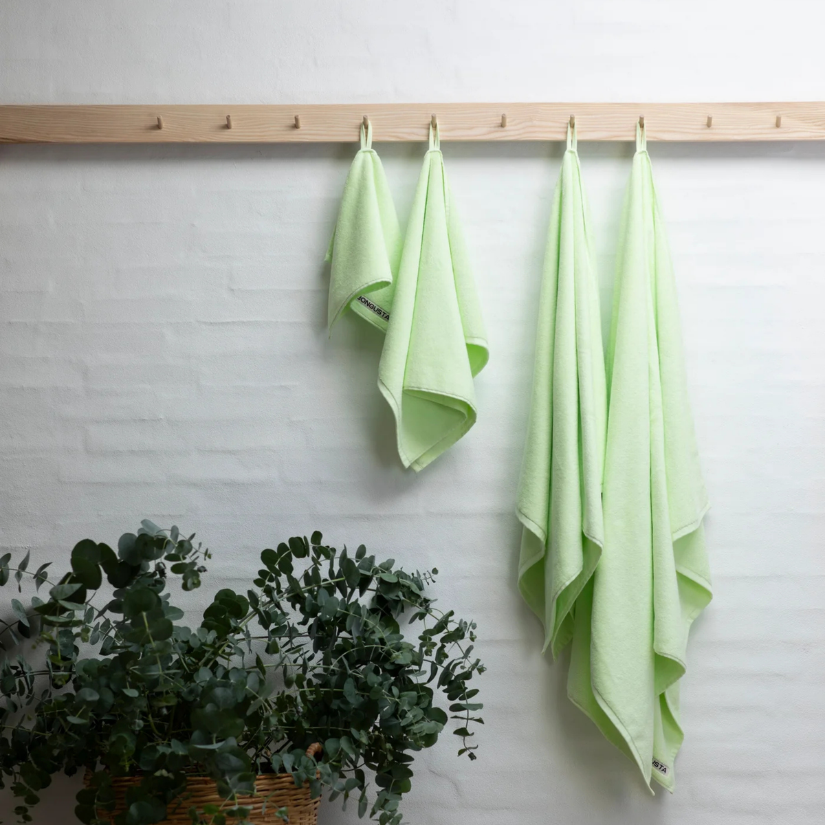 Vier Bongusta Naram Badhanddoek Lemon Sorbet 70/140 handdoeken hangen aan houten haringen tegen een witte bakstenen muur, met een mandje eucalyptus eronder.