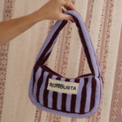 Een hand houdt de Bongusta Naram Mini Bag in Wine & Lavender Blue vast, met paarse en donkerrode strepen met een BONGUSTA-label tegen een achtergrond met kantpatroon.