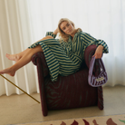Een vrouw leunt zijwaarts op een fauteuil met kastanjebruin patroon en houdt de Bongusta Naram Mini Bag Wine & Lavender Blue vast. Ze lijkt ontspannen in een zacht verlichte kamer met witte gordijnen achter haar.