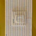 Drie beige handdoeken met blauwe strepen uit de Bongusta Naram collectie, elk met het label BONGUSTA, zijn gestapeld op een mosterdgeel oppervlak. Ze zijn gemaakt van biologisch katoen en passen perfect bij de Bongusta Naram Badmat Moonbeam & Ultramarine 50/80.