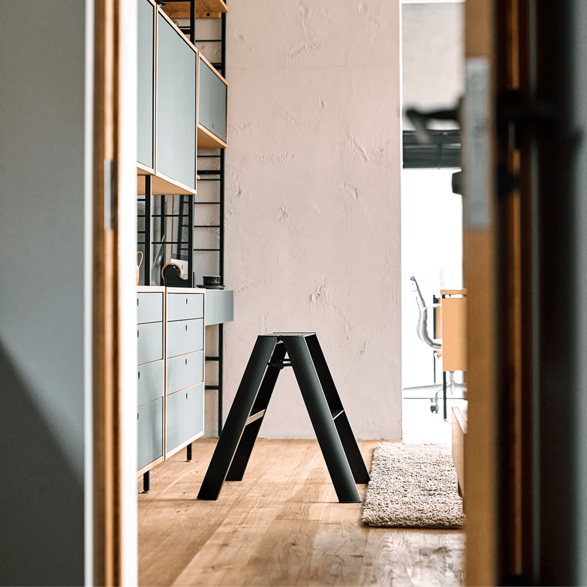 Een modern kantoor met houten vloeren, de Hasegawa Lucano Ladder 2 Step Zwart van Hasegawa in het midden, ingebouwde planken met kasten aan de linkerkant en een vergaderruimte op de achtergrond - allemaal subtiel geïnspireerd op Japans design.